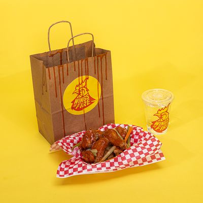 A bag with a sticker for a fake brand called Wings of Fire next to a basket of chicken wings and a clear plastic cup with another sticker for Wings of Fire on a yellow backdrop.