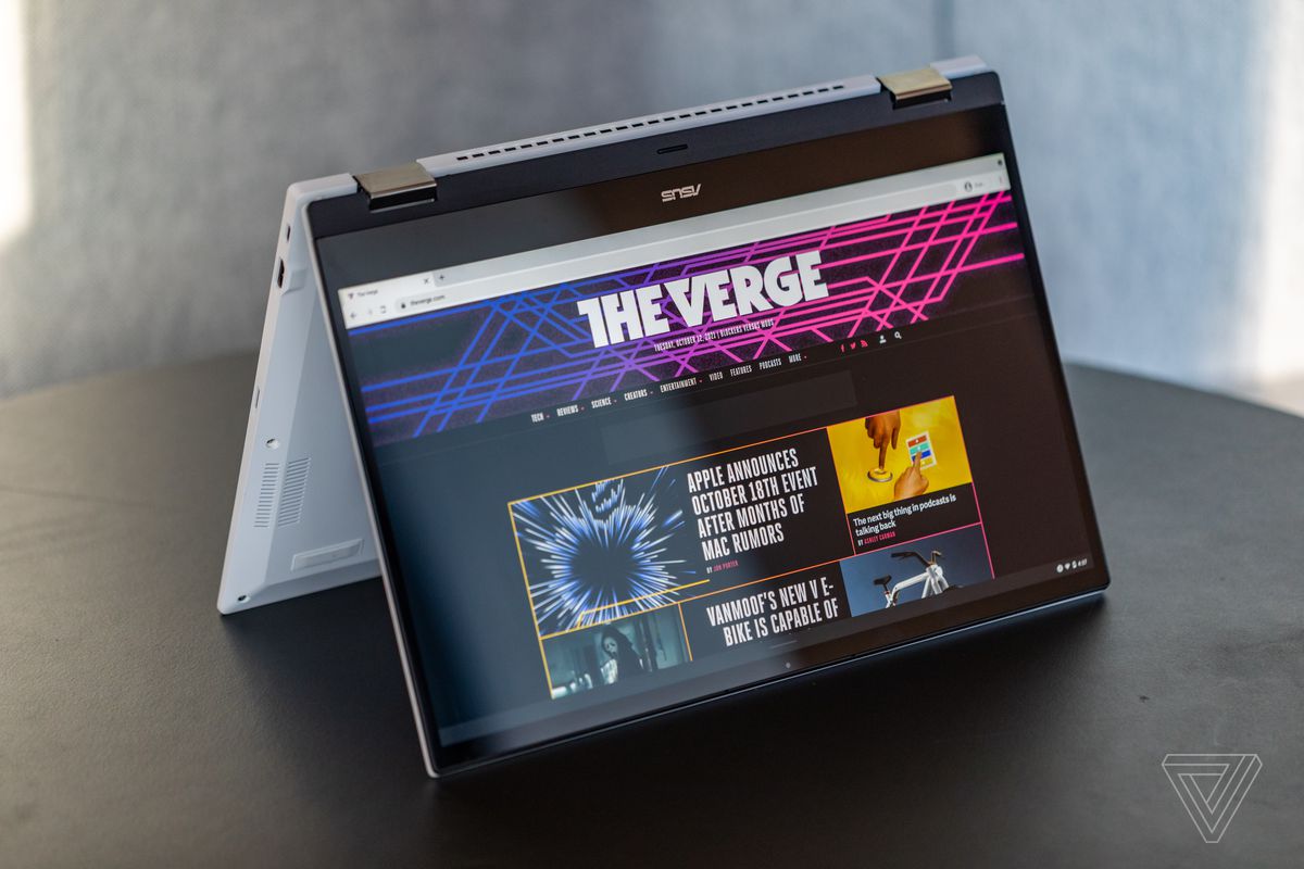 The Asus Chromebook Flip CX5 in tent mode, angled to the left on a black table. The screen displays The Verge homepage.