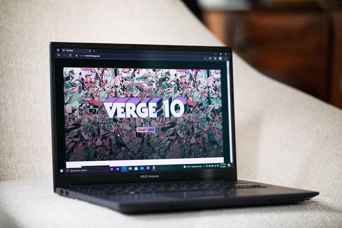 The Asus Vivobook Pro 14 on a white plush chair open angled to the left. The screen displays The Verge homepage.