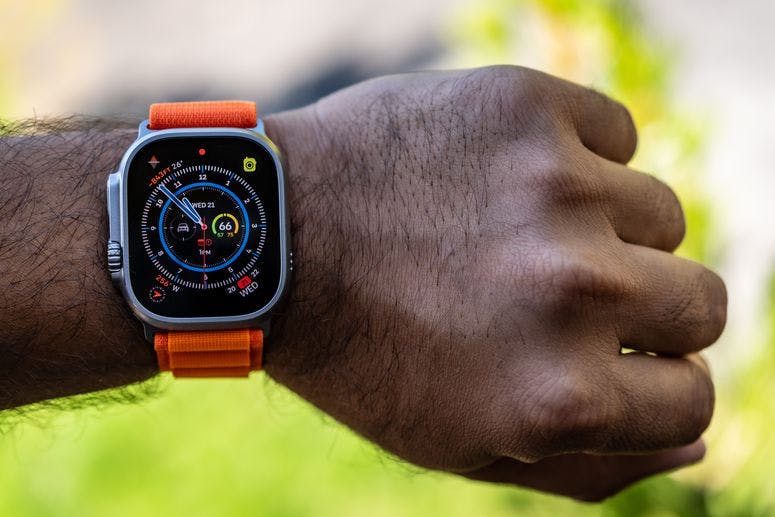 An Apple Watch Ultra with orange Alpine Loop strap on the left wrist of an adult man, with blurred green background.