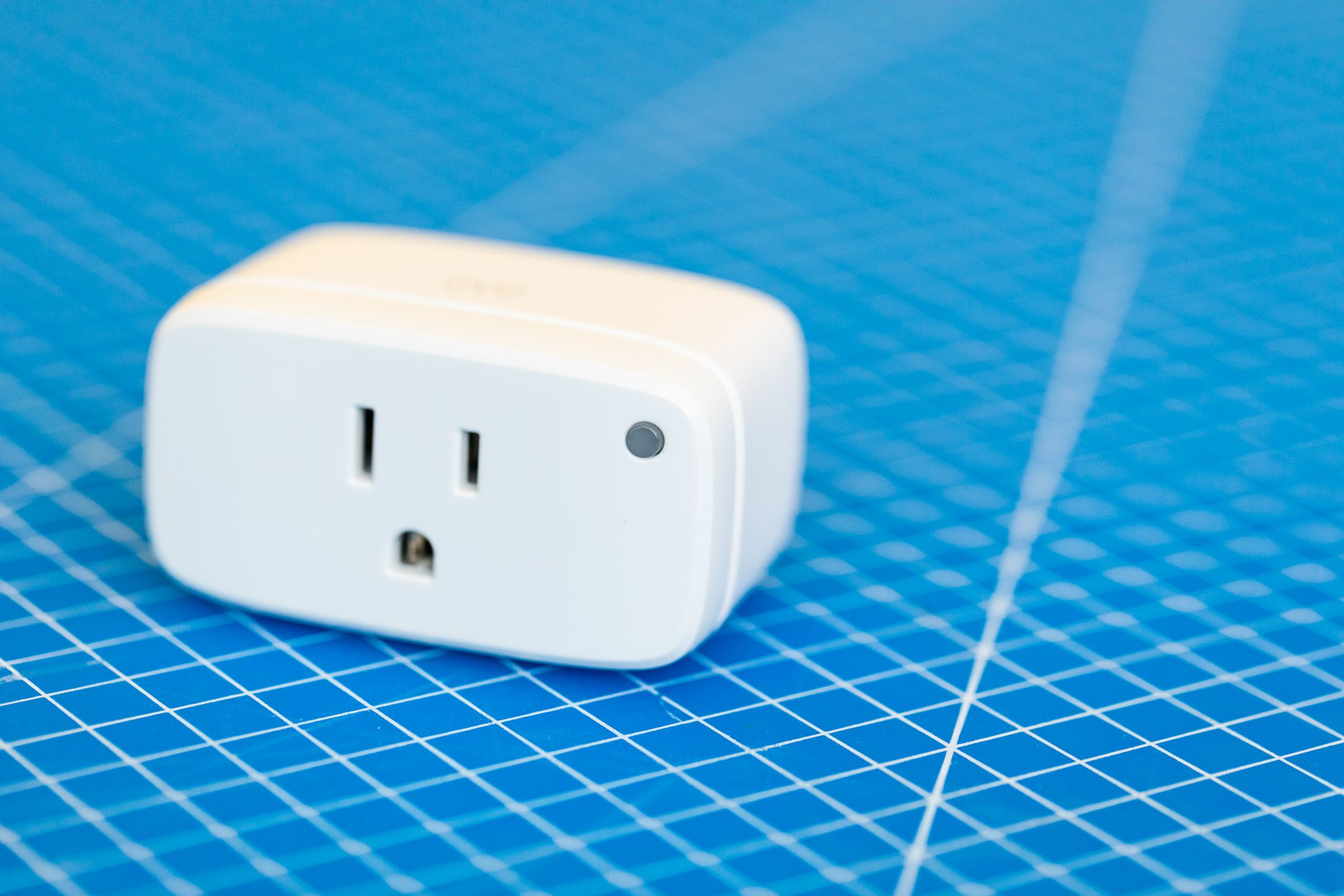 Eve Energy smart plug. A white rectangular prism with the broad face front. The corners and edges are rounded, there is a three-prong outlet on the front and a green LED on the upper-right corner of the front face. It sits on a blue background with a grid of white lines.